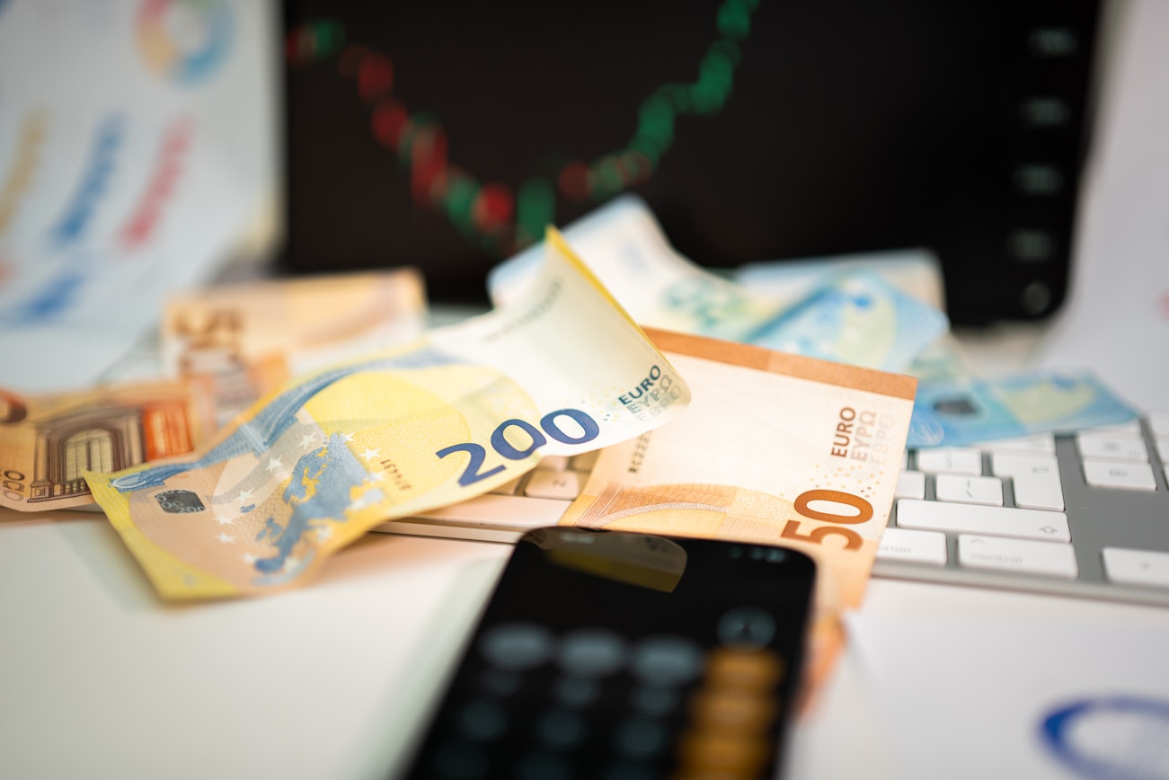 Euro banknotes on a keyboard with a phone calculator and a trading chart on a monitor in the background.