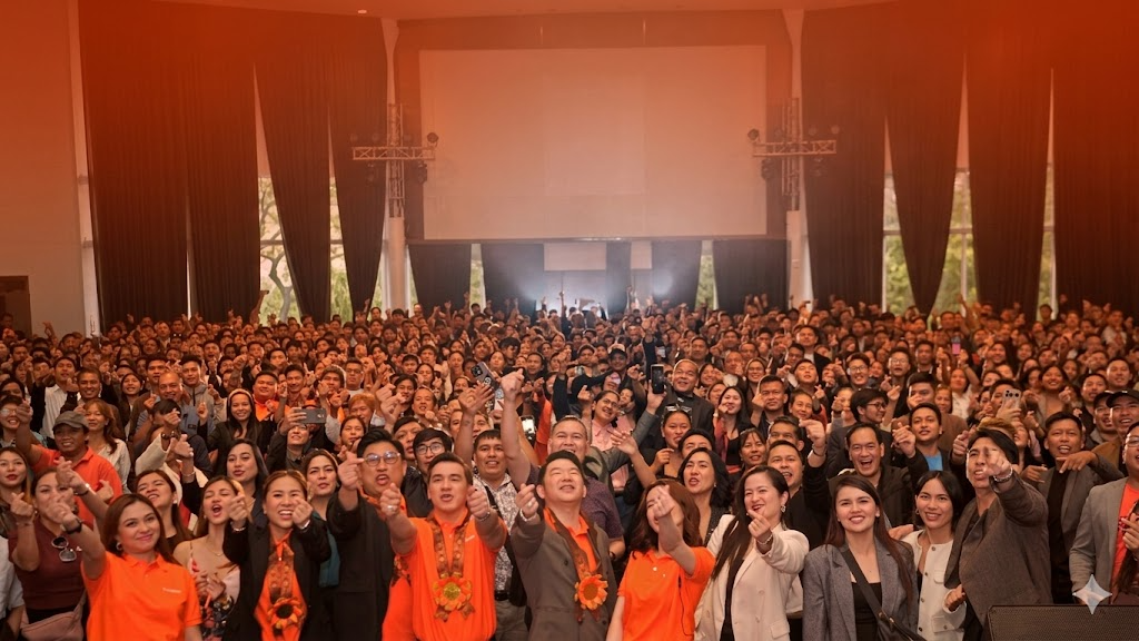 Large gathering of Ascendra members in an auditorium, many wearing orange and celebrating together.