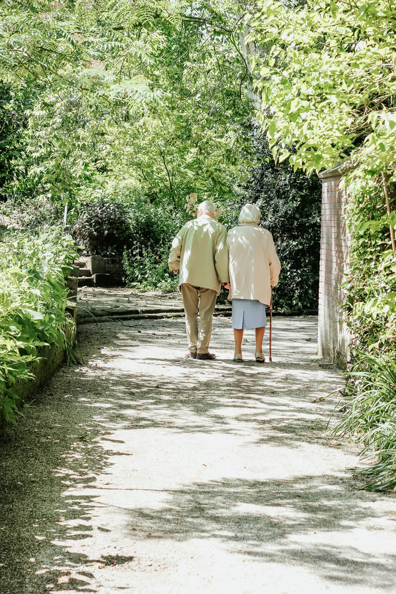 Elderly couple walking together on a sunlit garden path.
